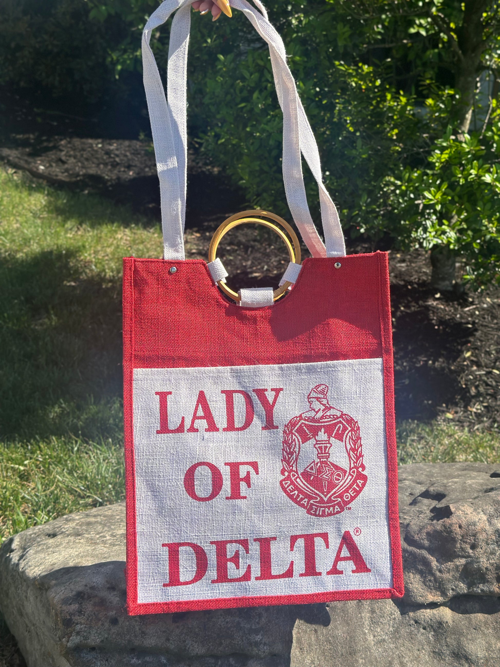 Red and white tote bag with 'Lady of Delta' text and logo on a stone surface.