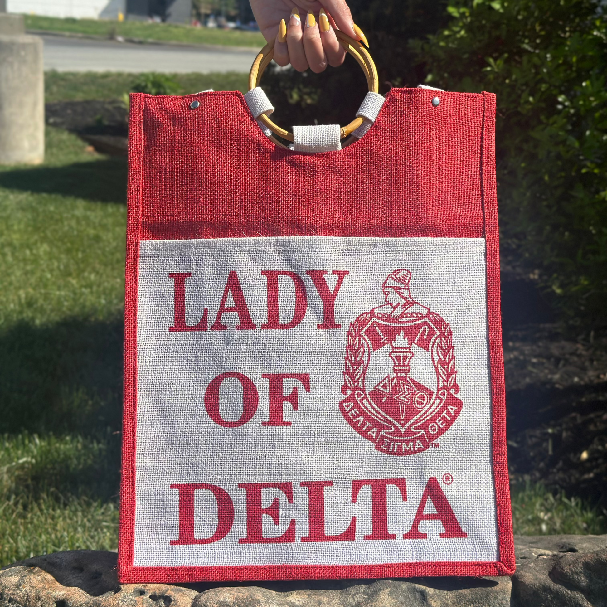 Red and white tote bag with 'Lady of Delta' text and logo held by a hand outdoors.